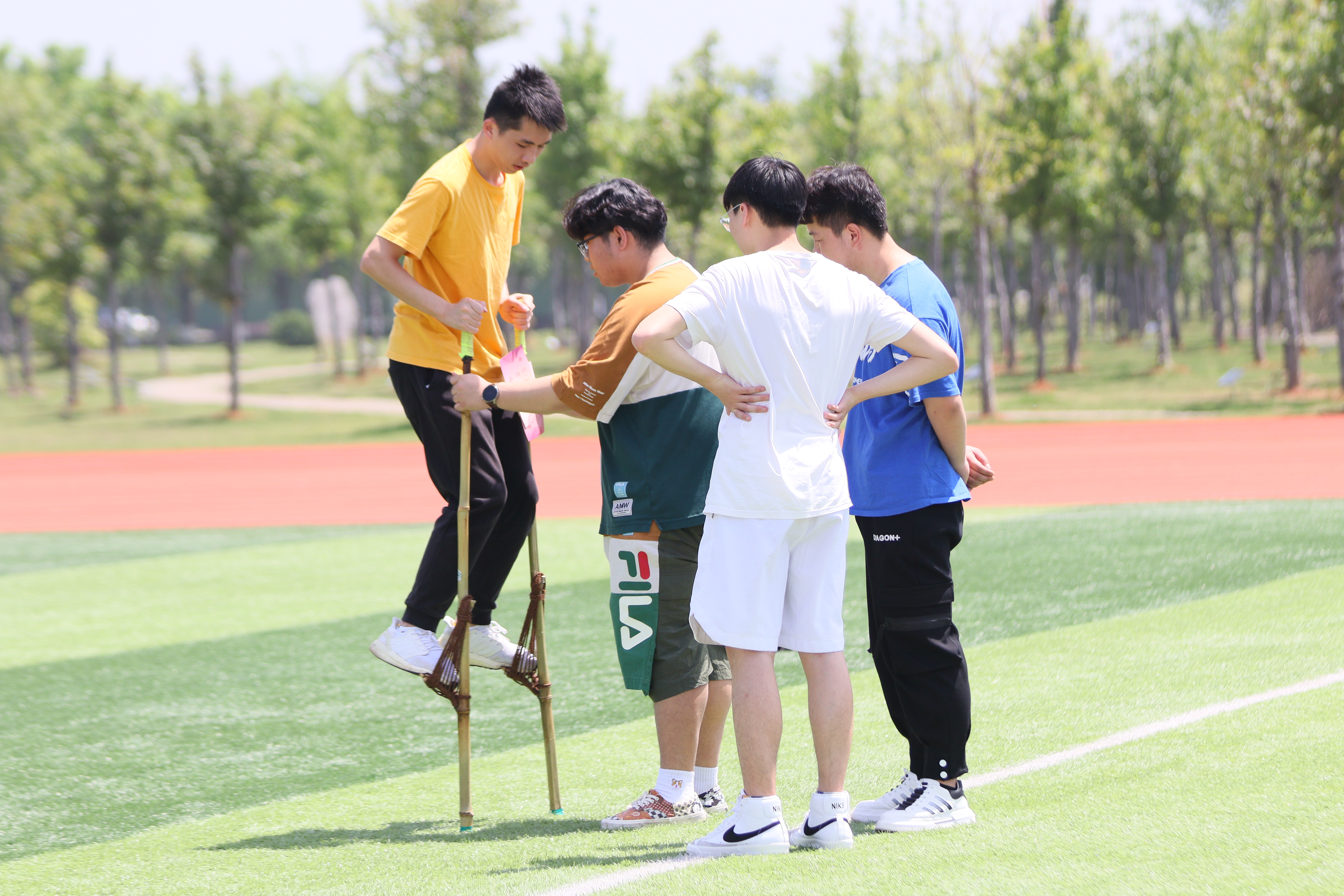 在草坪的一角，同学们正兴致勃勃地体验传统民俗活动——踩高跷，体验者双手紧紧地握住木杆，脚踩踏板，在工作人员的帮助下稳稳站住，紧张而又刺激地准备迈出第一步。