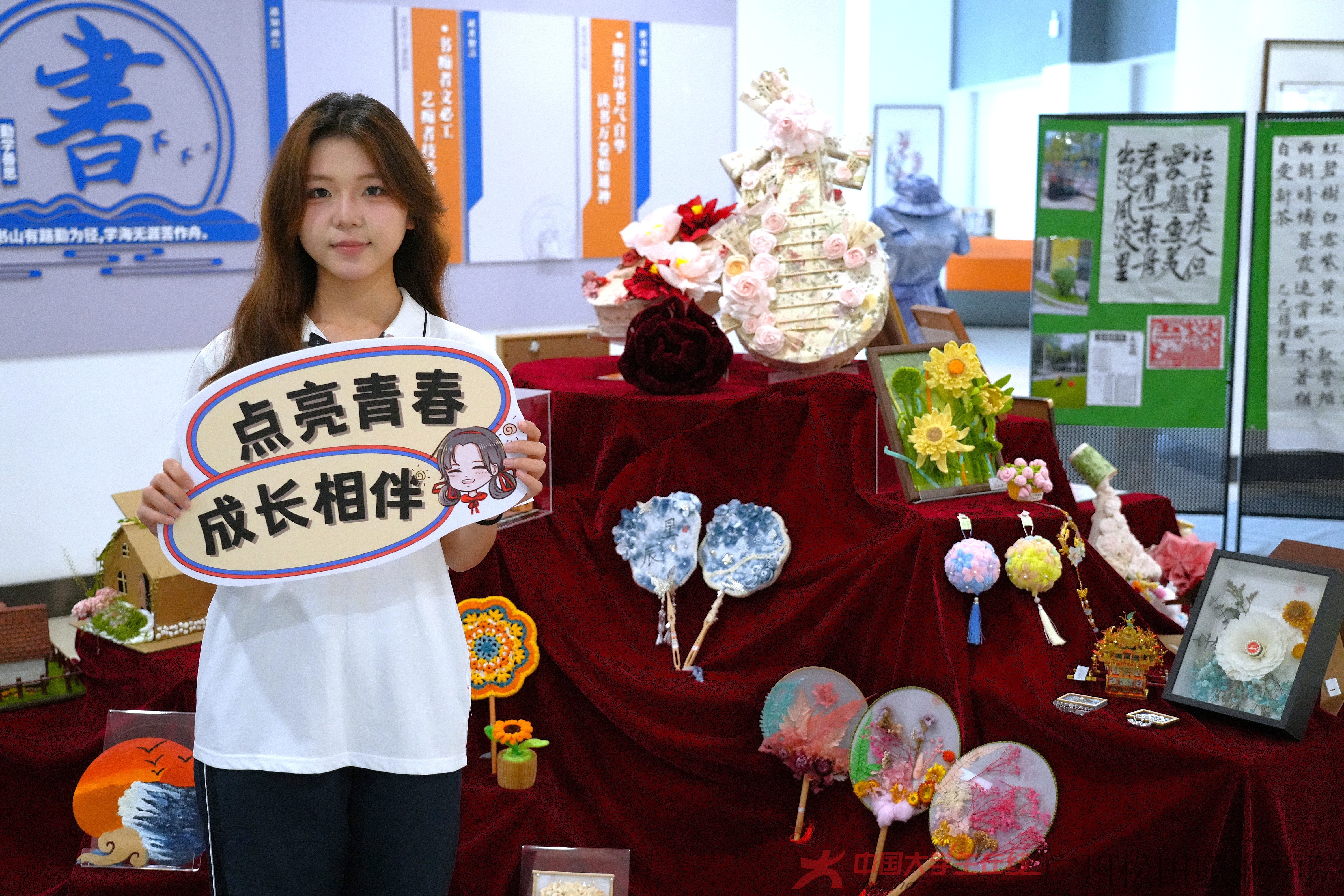 在勞動展現場，琳瑯滿目的手工藝品陳列于紅布之上，有古風團扇、創意花藝等。胡寶欣同學手持“點亮青春 成長相伴”手牌，笑容燦爛，為展覽增添青春活力與朝氣 。