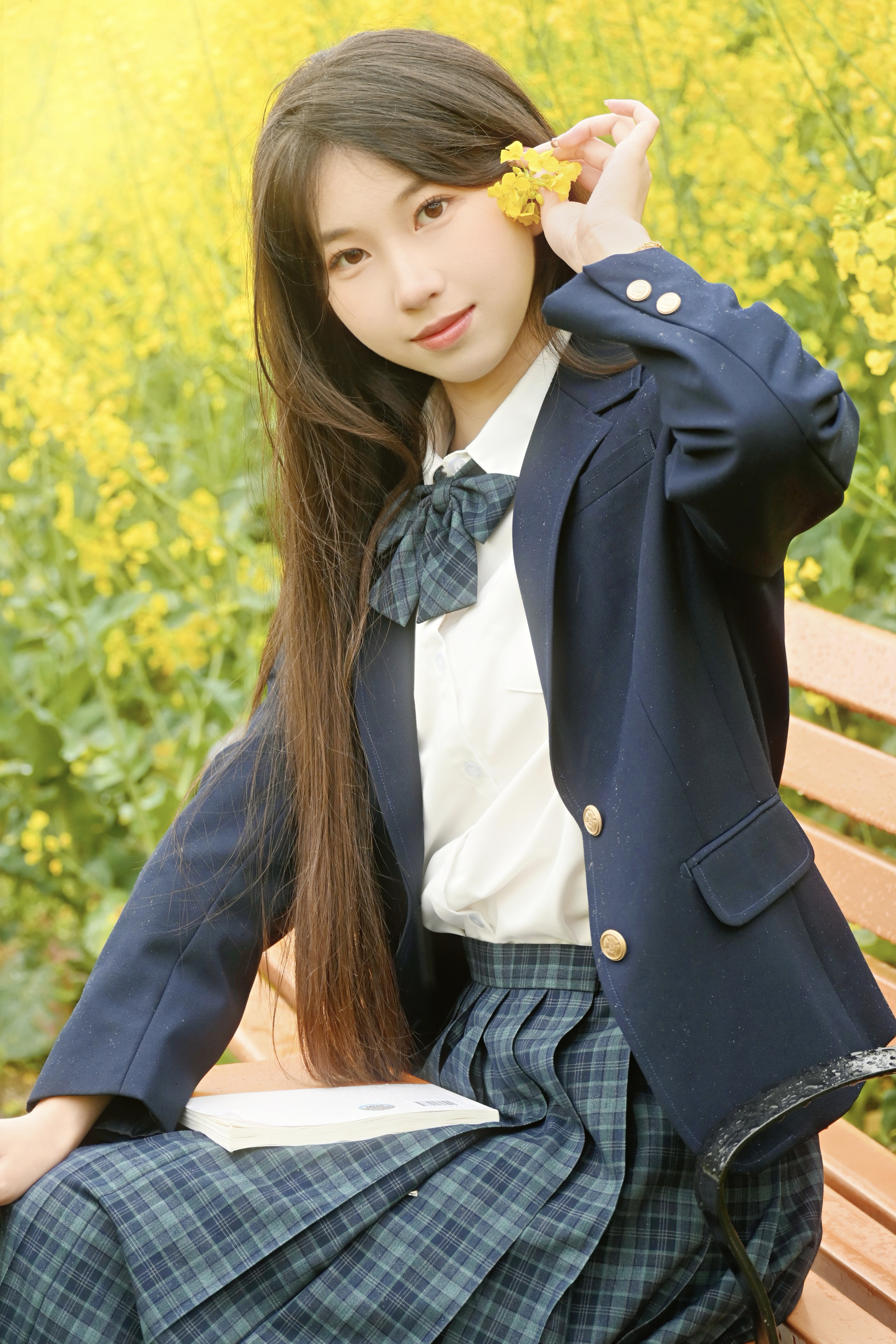 花海青春：阳光倾洒在无边的油菜花海，女孩身着清新制服坐在长椅上，手中轻轻拿着一朵黄色小花，眼神明亮，笑容甜美。盛雅婷/摄