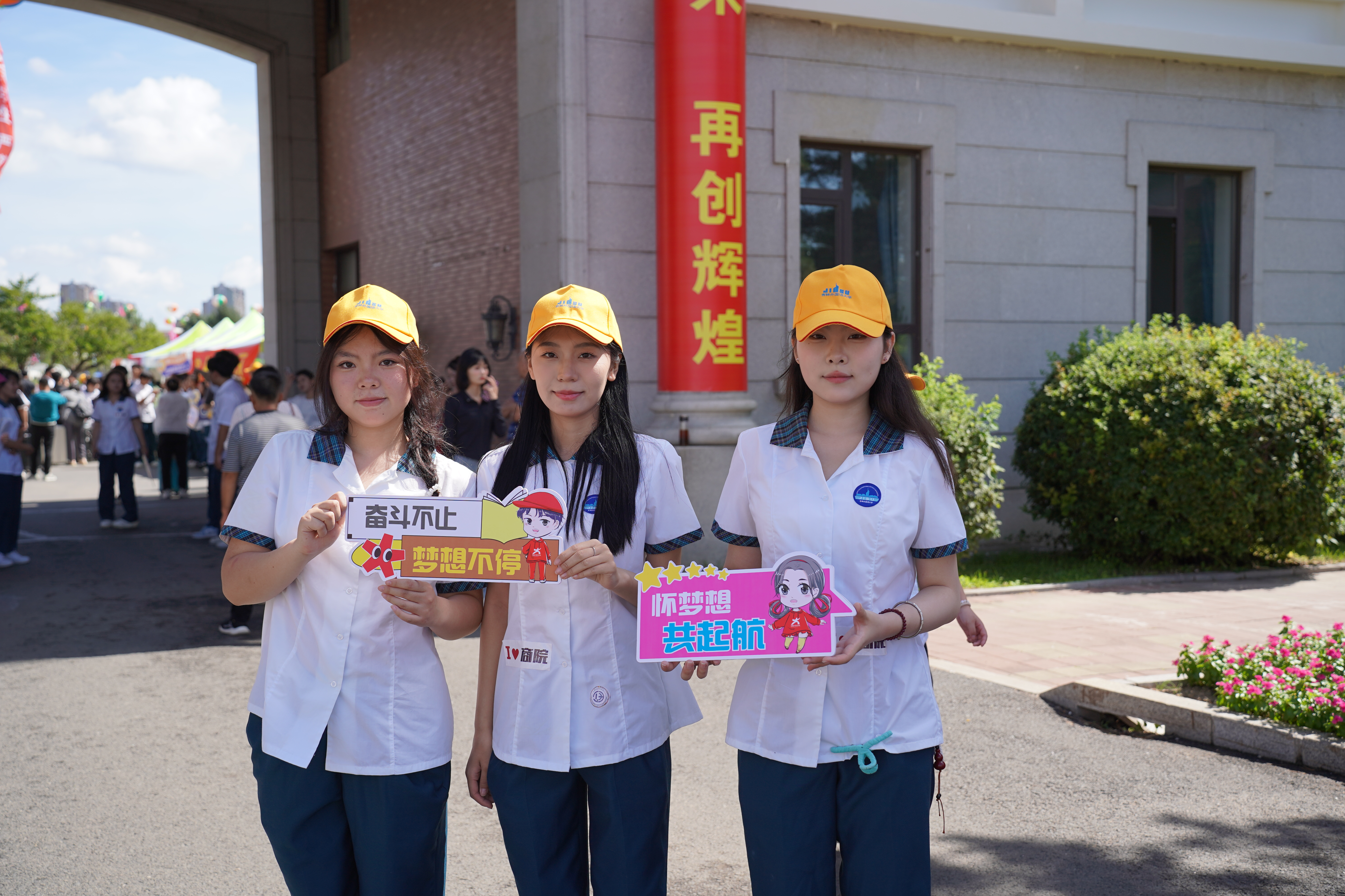 三位穿着校服的学姐手持迎新标语牌，在拱门大楼前