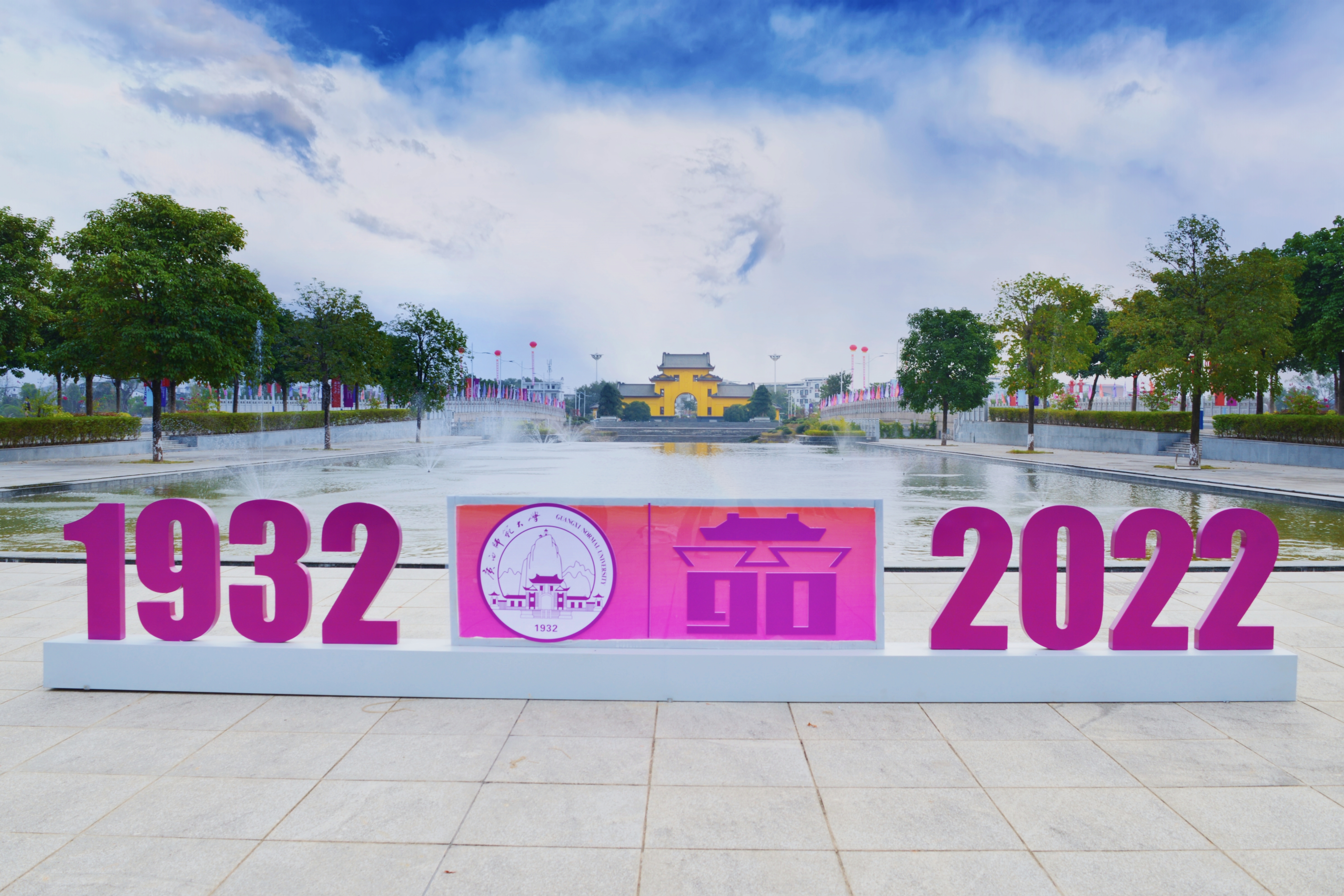 叹日月之不居，喜书香之绵延。九十年风雨同舟，一朝华诞祥瑞溢云霄。从最初的小小方圆，到如今的三大校区，广西师范大学在九十年的时光里走过了不凡的征程。校庆日当天，雁山校区东大门洋溢着喜庆的气息。“1932-2022”的标识与远处参考王城大门建造的校门遥相呼应，昭示着师大九十年以来依旧坚守初心、蓬勃发展。水池里的喷泉绽开朵朵水花，两旁道路上的彩旗和气球渲染着校园的活力，宣告着庆典的到来。.jpg